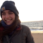 Smiling person with dark hair wears a Carhartt beanie on the beach.