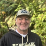 Ryan is smiling and wearing a baseball cap and a black sweatshirt as he poses in front of trees 