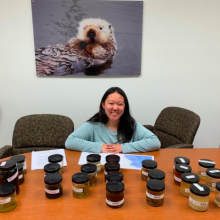 Student in aqua blue shirt sits at a table with several labeled specimen jars of various sizes. There is a photo of an otter on the wall.