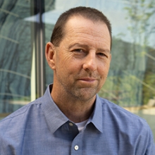 Man wearing blue shirt with close cropped dark hair stands in front of window.