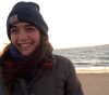 Smiling person with dark hair wears a Carhartt beanie on the beach.