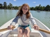 Smiling student with long reddish hair wearing a gray pullover hoodie and turquoise shorts rows a boat across pale green water. Deciduous trees in the background.