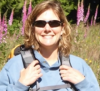 Susie Dunham is wearing sunglasses and a backpack while smiling in a field of lupins 